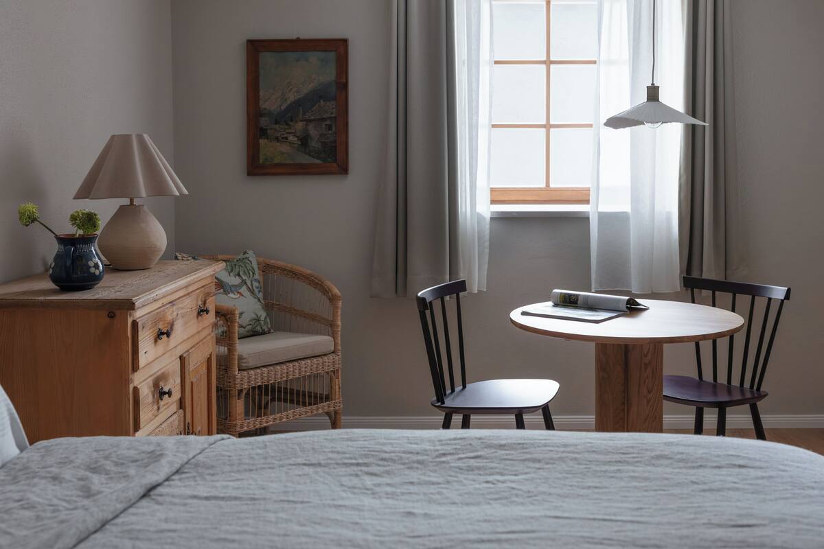 A bedroom with a bed, a wooden dresser, a wicker chair, a round table with two chairs, a lamp, and a painting on the wall.