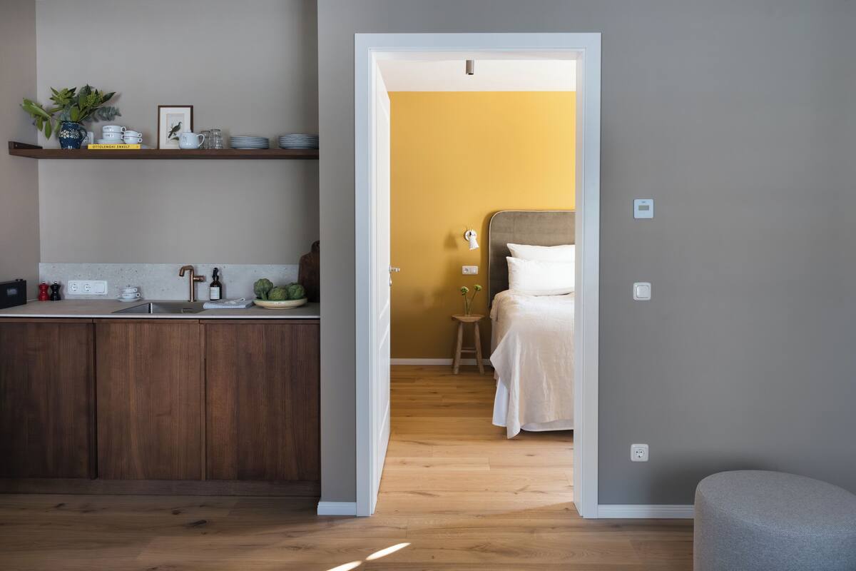 A modern kitchen with wooden cabinets and a built-in stove, leading to a bedroom with a bed and bedside table.