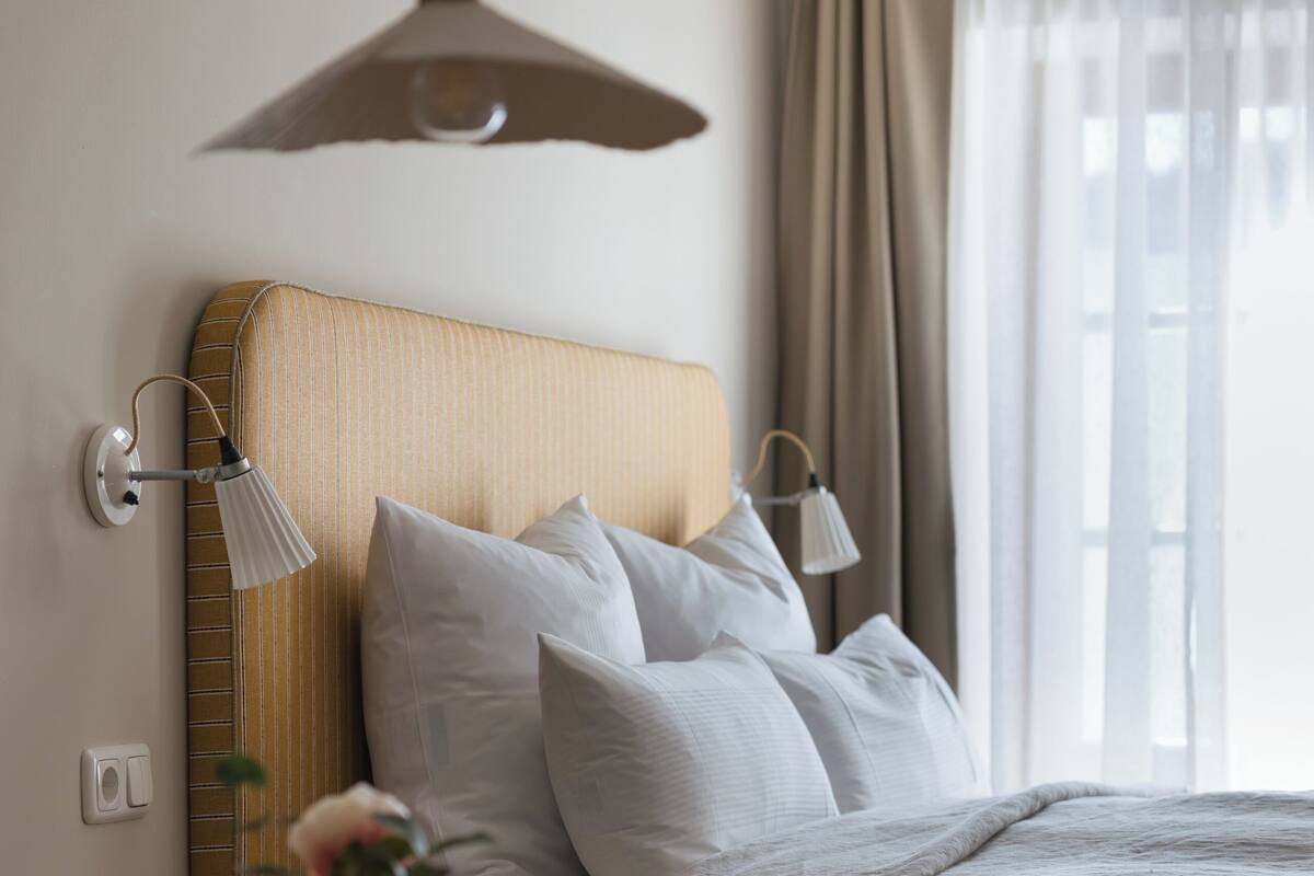A neatly arranged bedroom with a bed, bedside lamps, a bedside table with a vase, and a window with curtains.