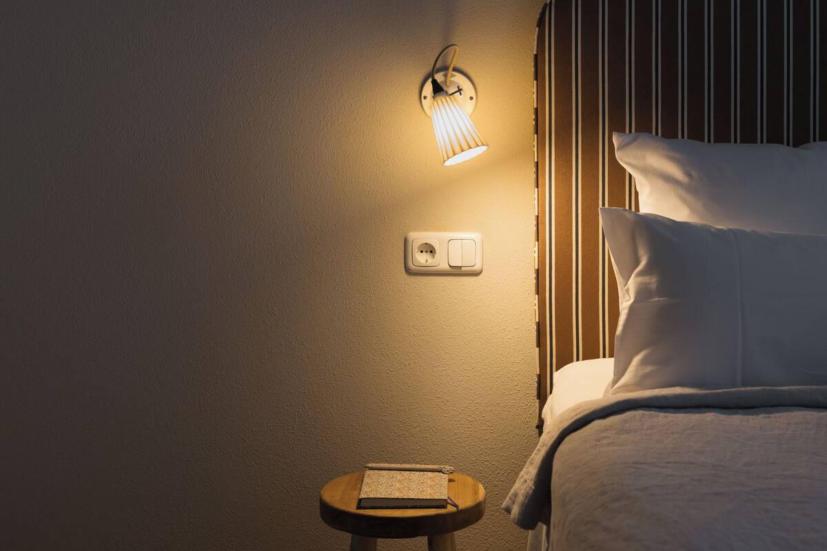 A dimly lit room with a bed, a small stool, and a wall-mounted light fixture.