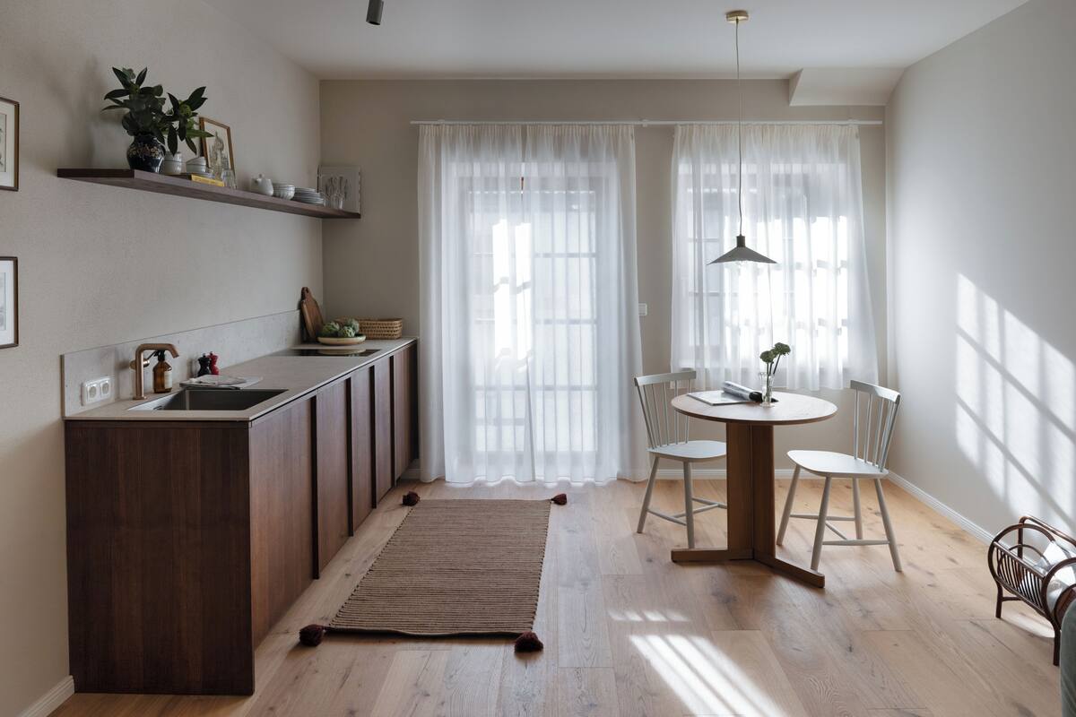 A modern kitchen with a wooden island, a dining area with a round table and chairs, and a large window with sheer curtains.