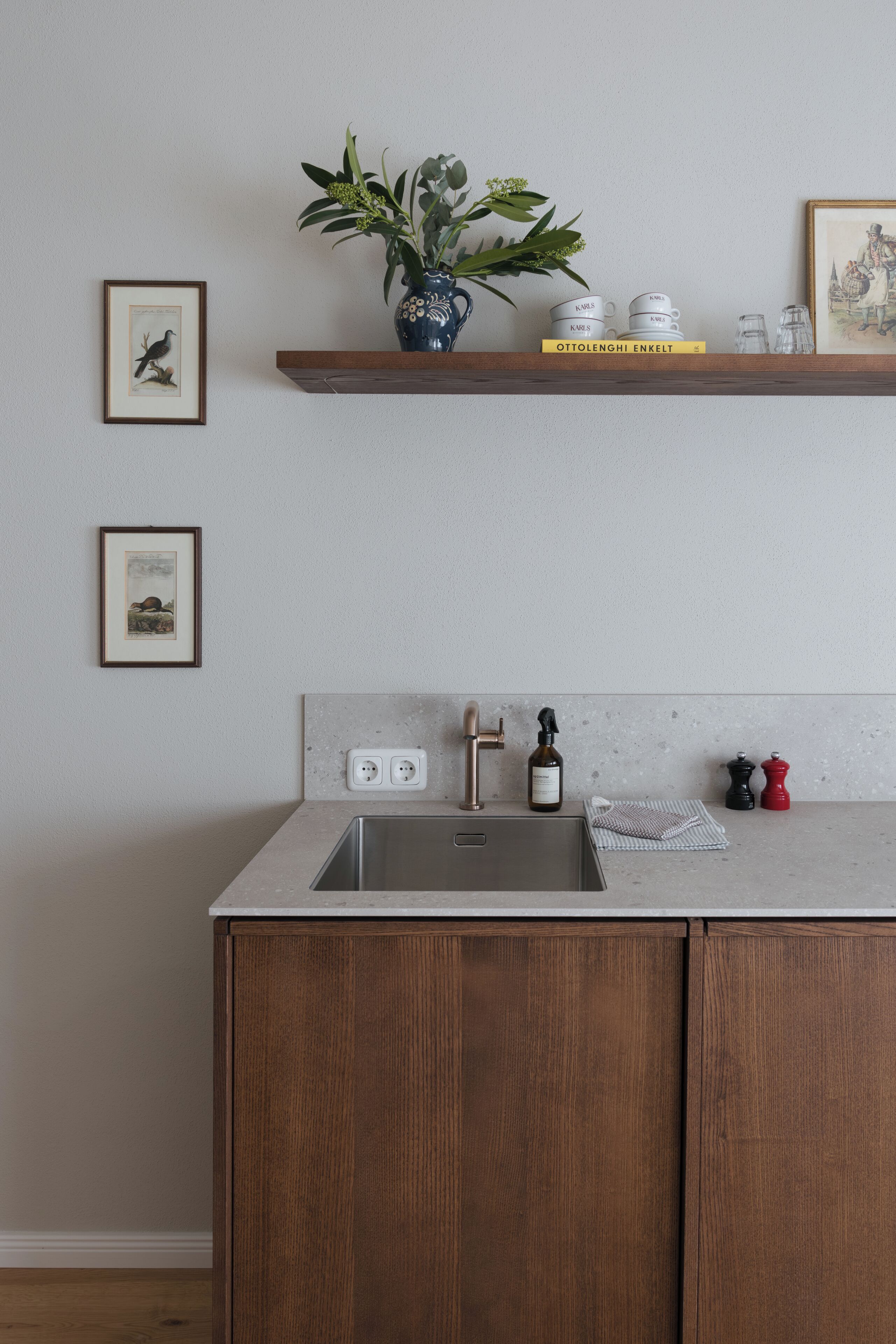 Una cucina con un armadietto in legno, un lavello e uno scaffale che contiene un vaso, libri e utensili da cucina.