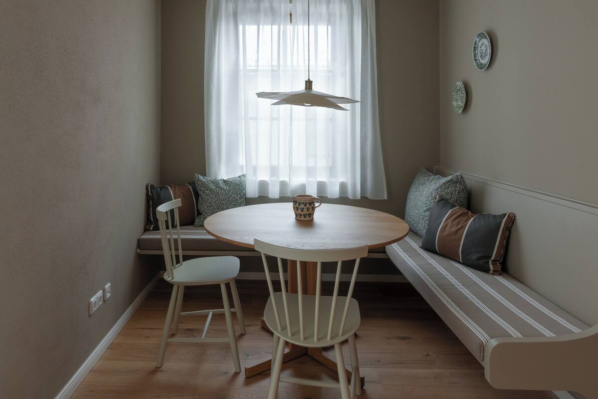 A cozy dining area with a round table, two white chairs, a bench with cushions, a hanging light fixture, and a window with sheer curtains.