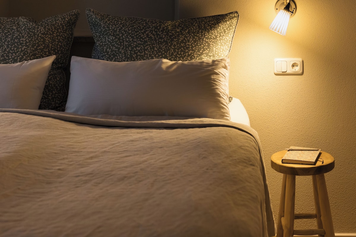 A bedroom with a bed, pillows, a nightstand, a lamp, and a power outlet.