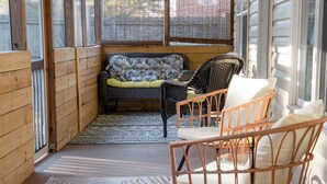 Un porche de madera con un sofá y sillones de mimbre, un tapete estampado y vistas a través de las ventanas.