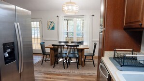 Una cocina con un refrigerador de acero inoxidable, una mesa de comedor de madera con sillas, una araña y una ventana con cortinas blancas.
