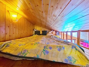 Ein gemütliches Schlafzimmer im Dachgeschoss mit Holzboden und -wänden, einer großen blumigen Tagesdecke und einem Nachttischchen mit Lampe.