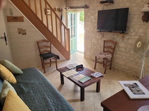 A cozy living room with a wooden staircase, a flat-screen TV, a fan, and a coffee table with books. - Charming Old House near Chinon with Private Courtyard (Tavant)