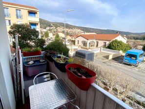 Ein Balkon mit Topfpflanzen, einem Metalltisch und Blick auf eine Wohngegend mit Häusern und einem blauen Lieferwagen.