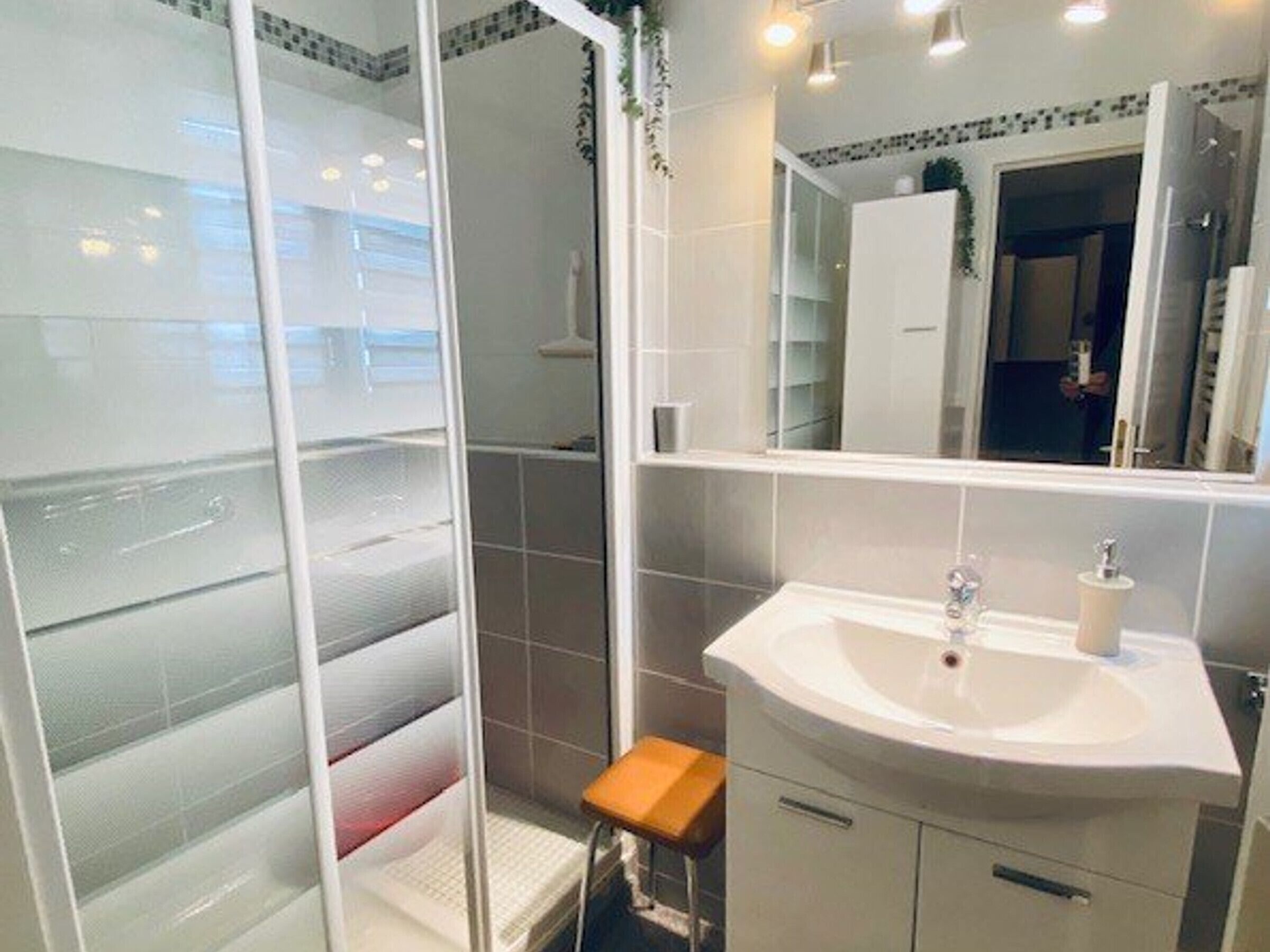 A bathroom with a glass shower enclosure, a white sink with a mirror above it, and a small wooden stool.