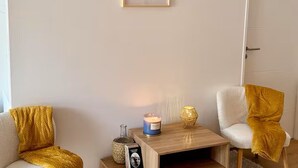 A room with a white wall, a framed picture of a coastal scene, a small wooden table with a candle, a bottle, and a book.