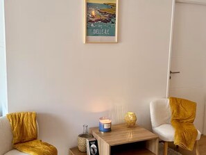 Ein Zimmer mit weißer Wand, einem gerahmten Bild einer Küstenlandschaft, einem kleinen Holztisch mit Kerze, einer Flasche und einem Buch.