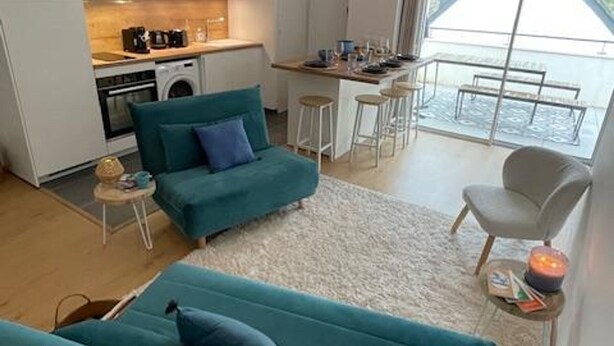 A modern living room with a teal sofa, a small table, and a view of the kitchen and balcony.