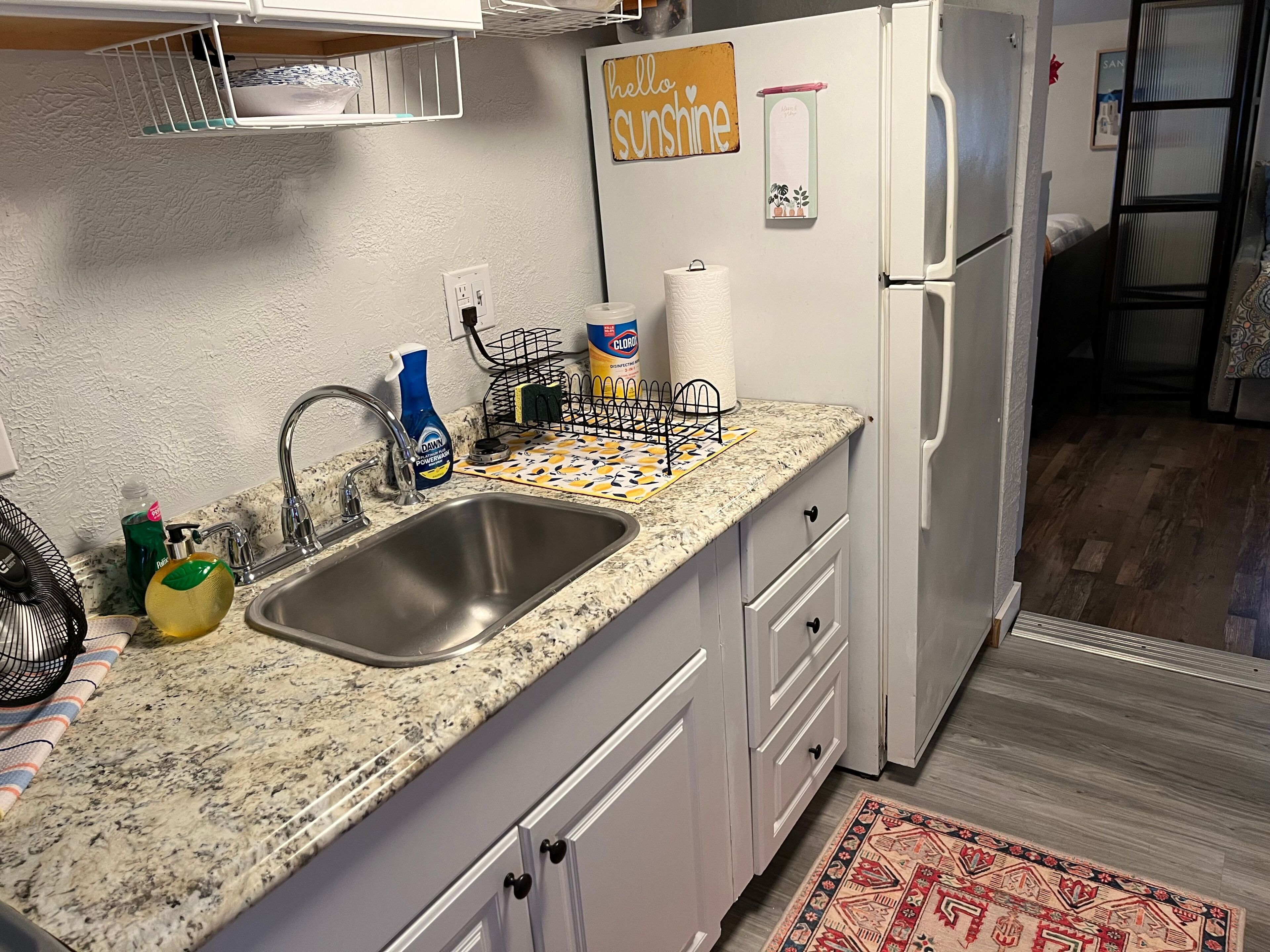 A kitchen with a granite countertop, stainless steel sink, white cabinets, and a refrigerator.