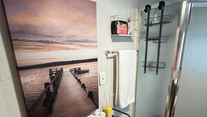 Un baño con ducha de vidrio, un perchero, un estante con artículos de aseo y un cuadro de atardecer en la pared.