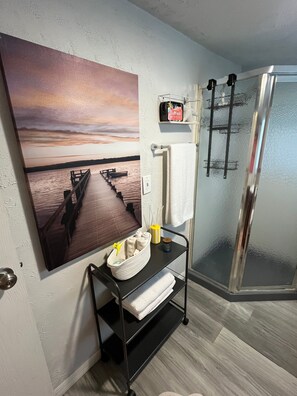 Une salle de bain avec une douche en verre, un porte-serviettes, une étagère avec des articles de toilette et un tableau représentant un coucher de soleil au mur.