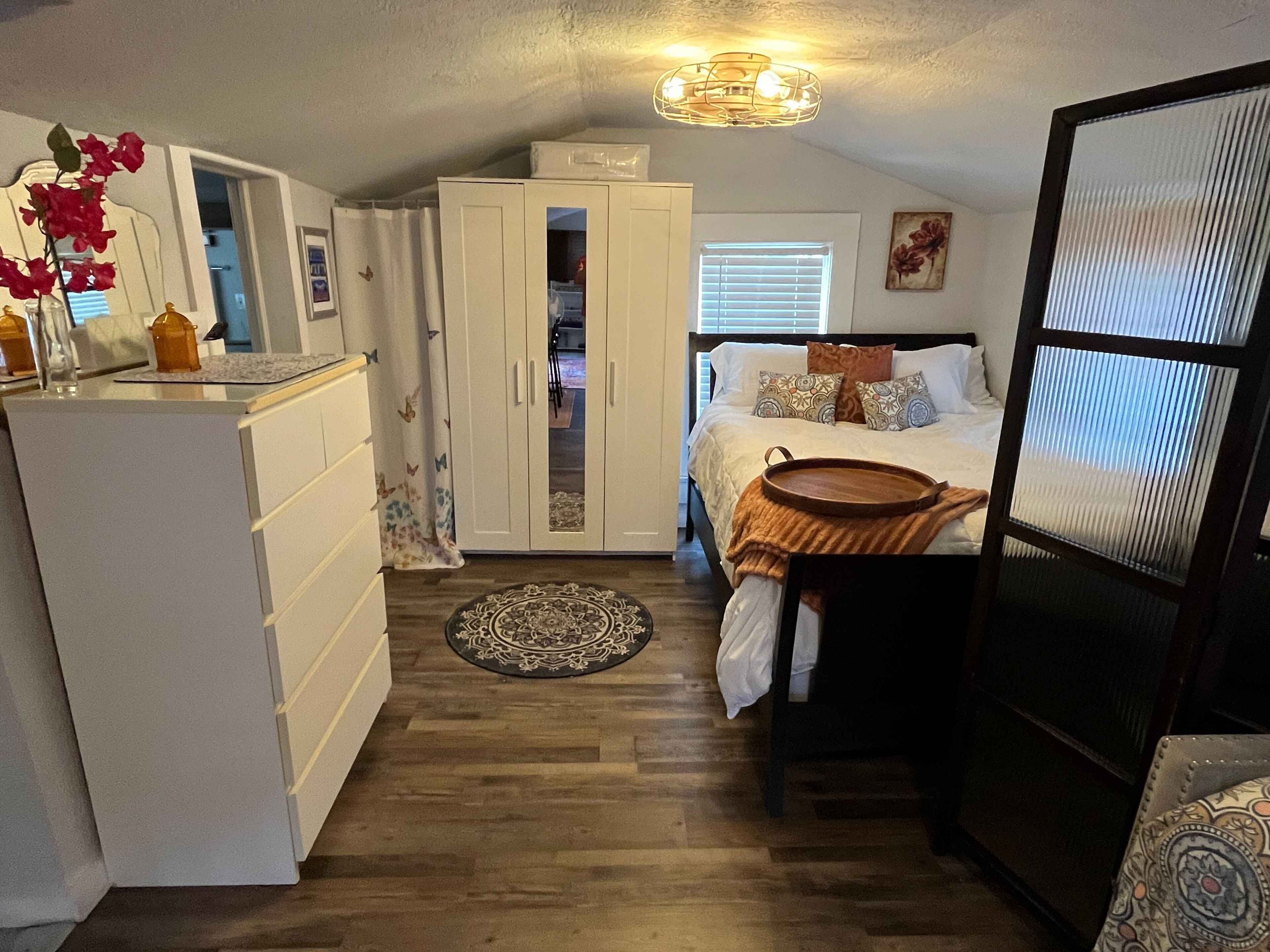 A compact bedroom with a bed, bedside table, wardrobe, and a small kitchen area with a refrigerator and a microwave.