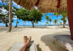 A beach scene with a thatched roof structure, palm trees, and a clear view of the ocean. - Beachfront 2bedRm 2Bath Apt G1-3 PlayaNuevaRomana w/Pool,JacuziSpa,GolfTenis,Gym (Playa Nueva Romana)