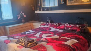 A bedroom with a large bed, a bedside table, a window with curtains, a wall-mounted shelf with decorative items, and a hanging light fixture. - La Casa di Sandhya (Udine)