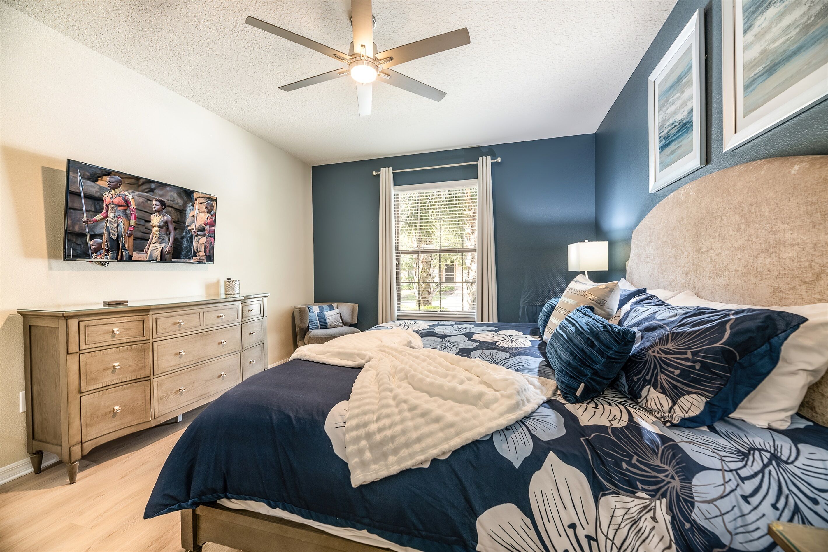A bedroom with a bed, dresser, ceiling fan, window, and artwork on the wall.