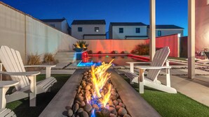 A backyard with a fire pit, two Adirondack chairs, a pool with red lighting, and a view of neighboring houses. - Shadow Stone | Private Pool & Spa | 16+ Guests! (Hurricane)