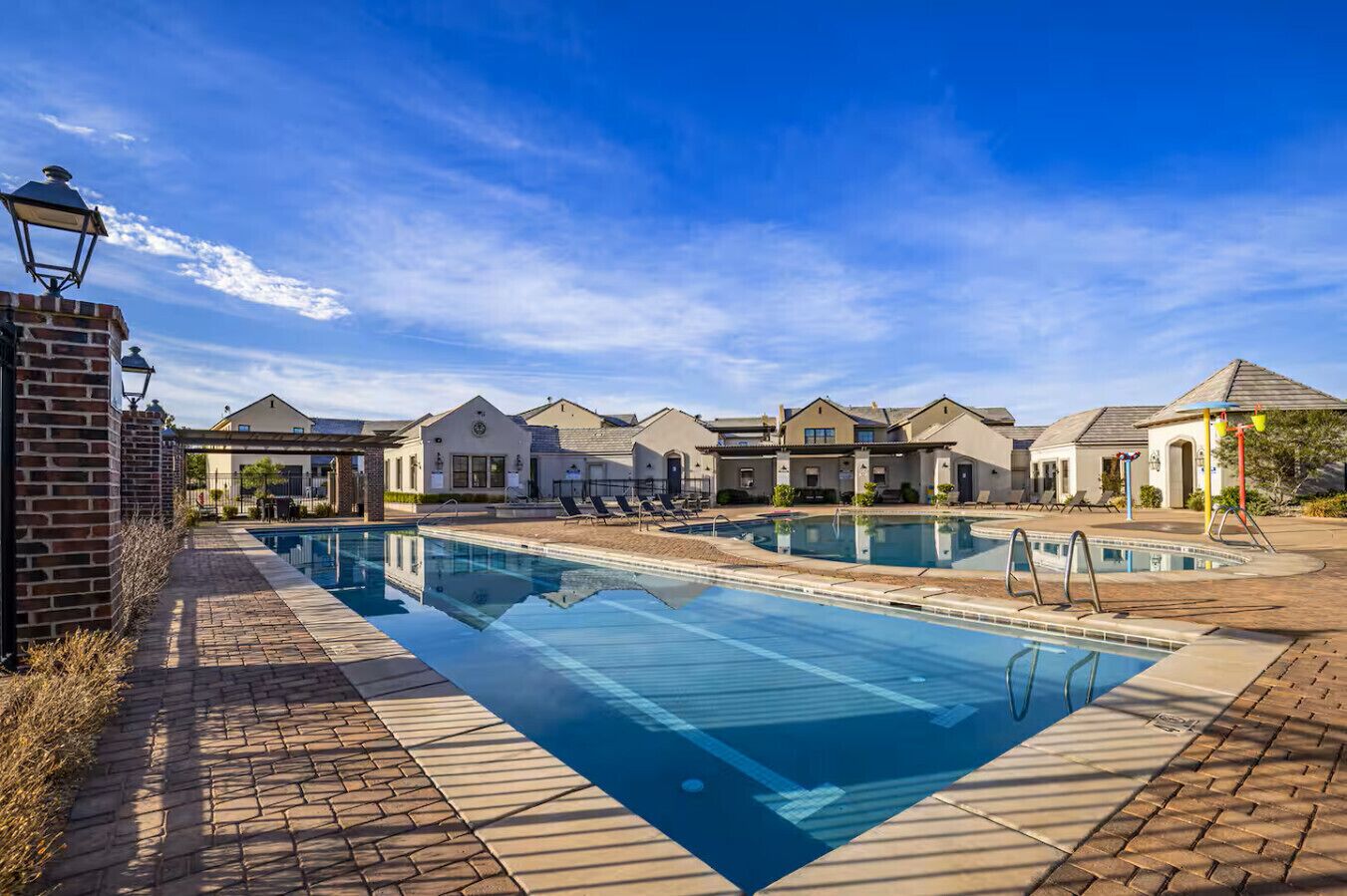 Una piscina con sillones de descanso, un sendero de ladrillos y una hilera de casas bajo un cielo despejado.