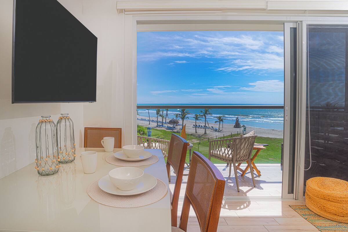 Un coin repas avec vue sur la plage, une table dressée avec des assiettes et des bols, et deux bouteilles en verre décorées de perles jaunes.