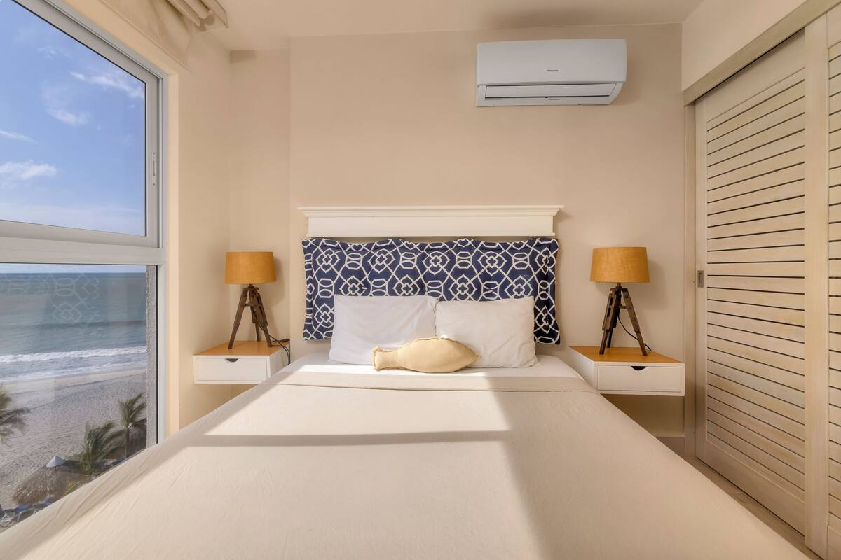 Une chambre avec une grande fenêtre donnant sur la plage, une tête de lit à motifs, deux tables de chevet avec des lampes et un appareil de climatisation.