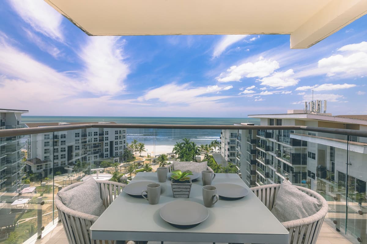 Un balcon avec une table dressée pour deux personnes, offrant une vue sur une plage et le paysage urbain.