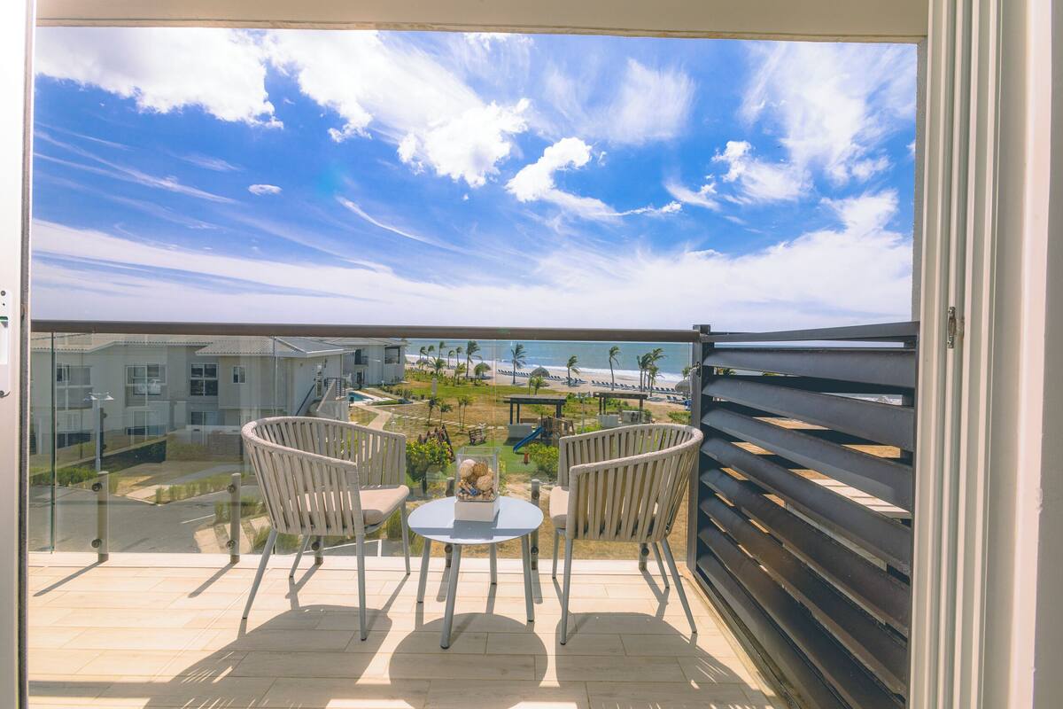 Un balcon avec une porte-fenêtre, deux chaises, une petite table et une vue sur une plage et des immeubles.