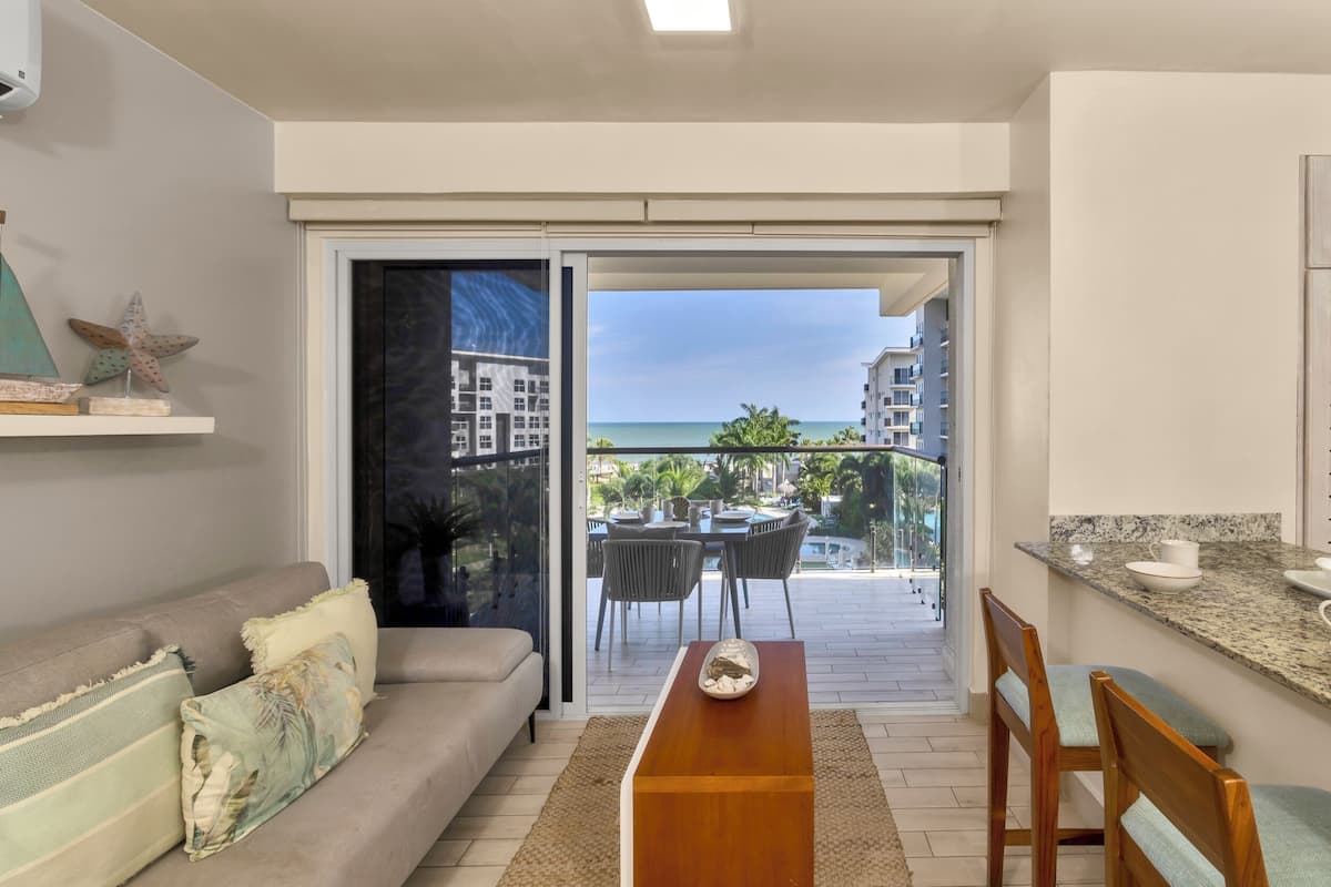 Un salon moderne comprenant un canapé, une table à manger et un coin cuisine. Une porte-fenêtre coulissante donne accès à un balcon offrant une vue sur l’océan et les bâtiments.
