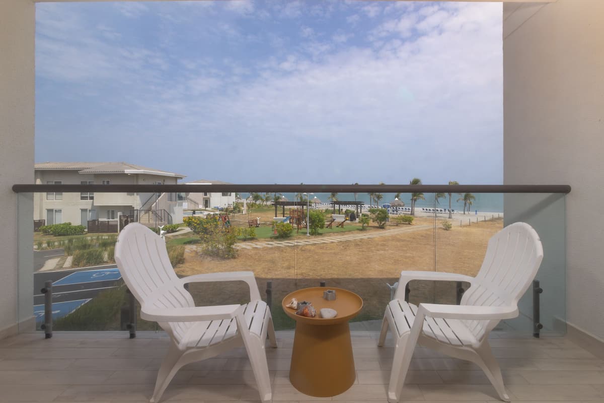 Un balcon avec deux chaises blanches et une petite table, donnant sur une plage et un terrain de jeux.