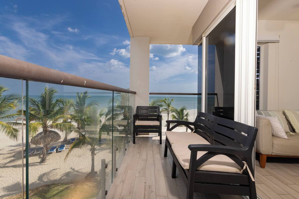 Un balcon doté d’une balustrade en verre, donnant sur une plage bordée de palmiers et offrant une vue sur un ciel dégagé.
