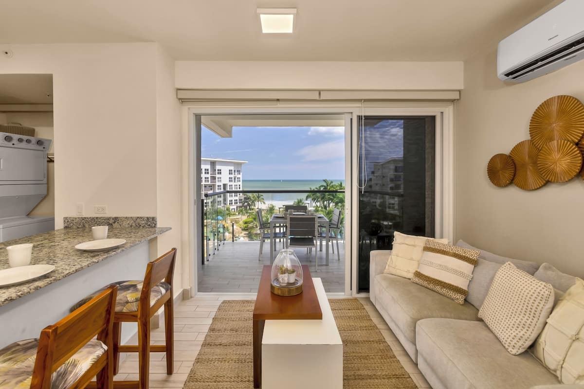 Une cuisine moderne avec des comptoirs en granit et un coin repas doté de chaises en bois. Une porte-fenêtre coulissante donne accès à un balcon offrant une vue sur l’océan et les bâtiments.