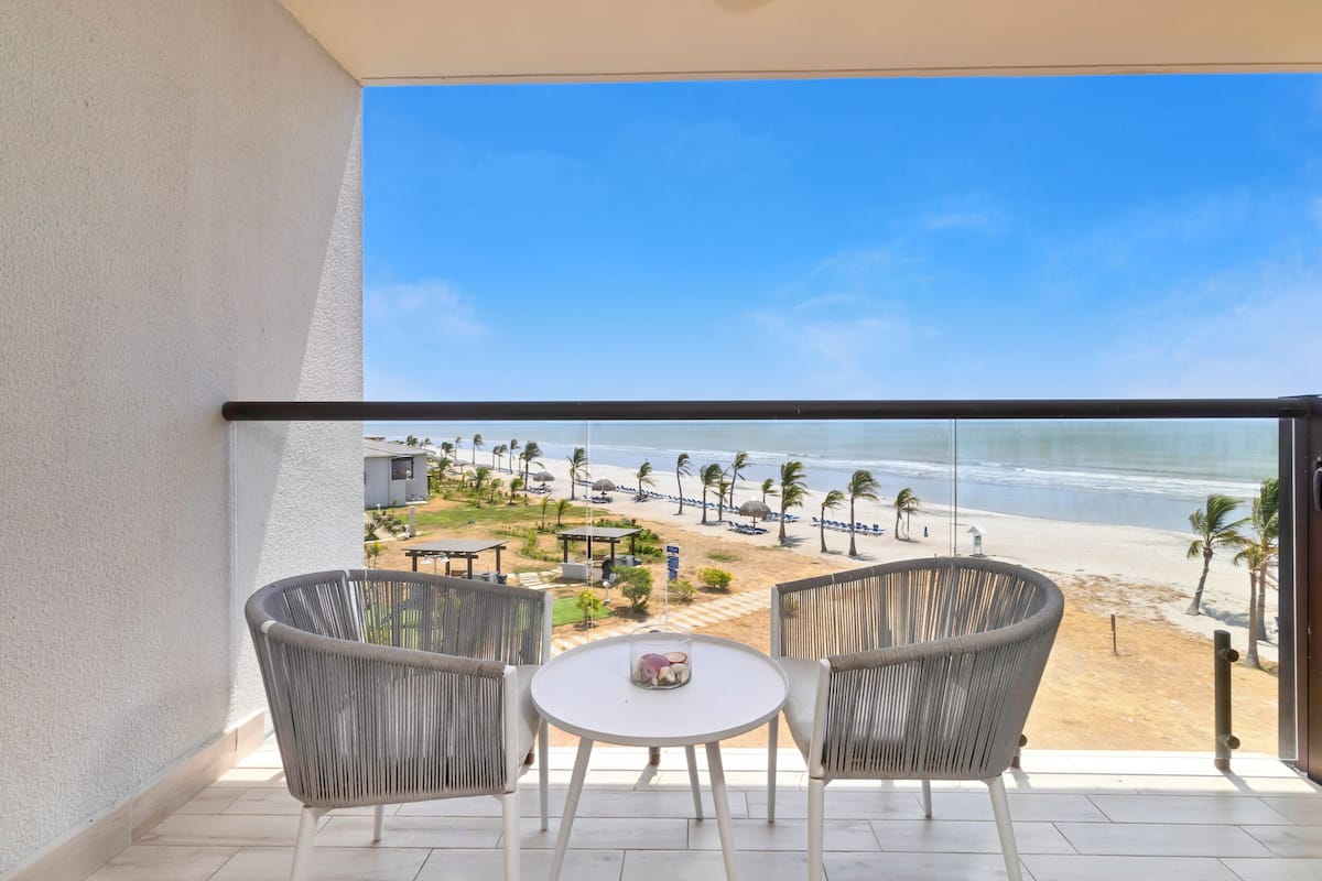 Un balcon avec deux fauteuils en osier et une petite table, donnant sur une plage bordée de palmiers et parsemée de parasols.