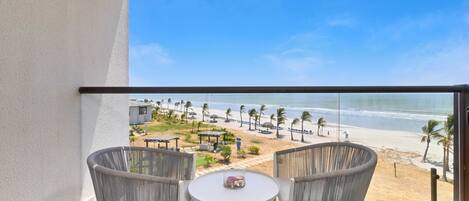 Ein Balkon mit zwei Korbstühlen und einem kleinen Tisch, mit Blick auf einen Strand mit Palmen und Sonnenschirmen.