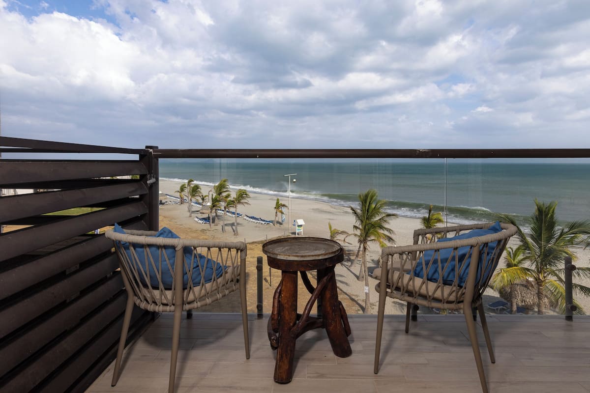 Un balcon avec des fauteuils en osier et une petite table, donnant sur une plage bordée de palmiers et sur l’océan.