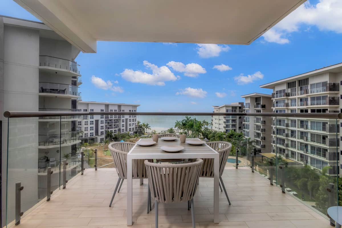 Un balcon avec une table et des chaises, donnant sur une plage et d’autres bâtiments.