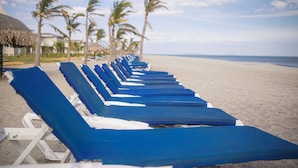 A row of blue lounge chairs on a sandy beach with palm trees and a thatched-roof structure in the background.