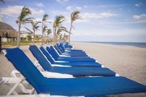 Una hilera de sillas reclinables azules sobre una playa arenosa, con palmeras y una estructura de techo de paja al fondo.