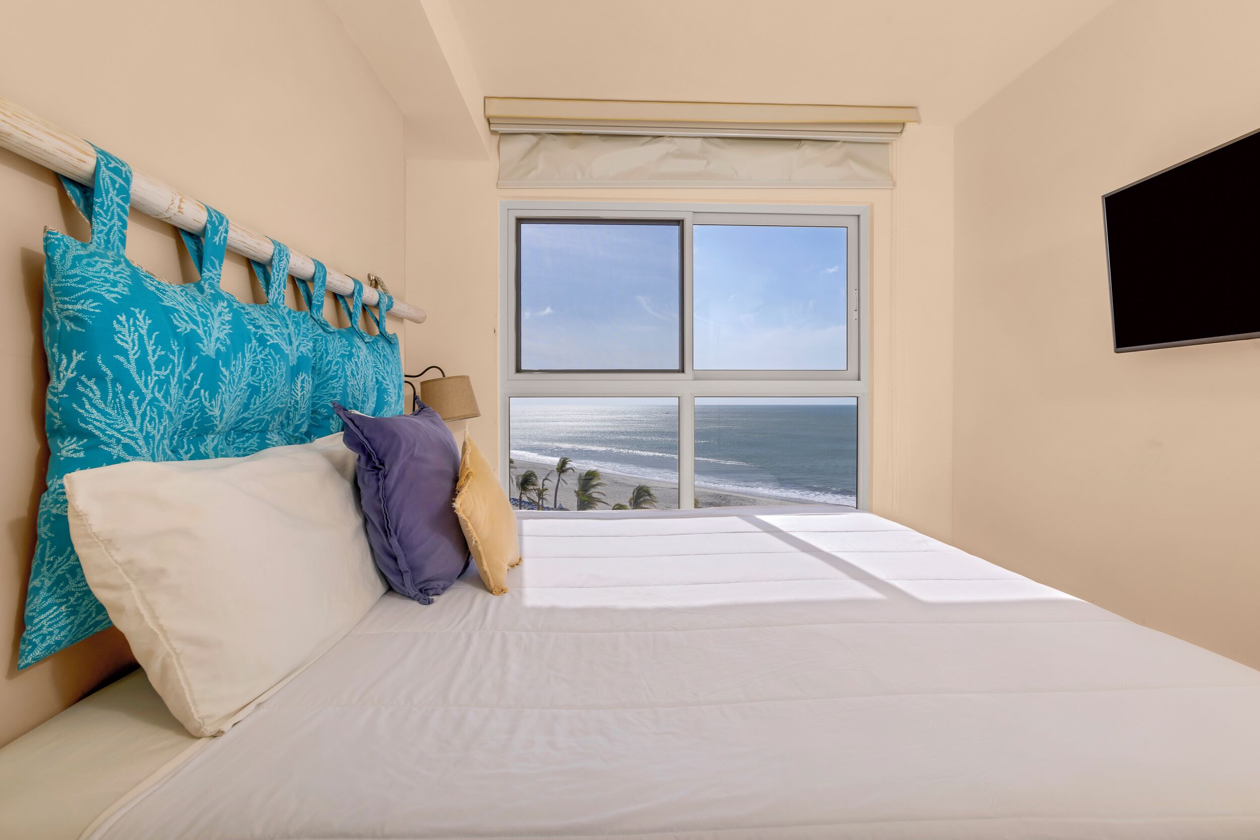 Een slaapkamer met een groot raam met uitzicht op de oceaan, een bed met wit beddengoed en een aan de muur bevestigde televisie.