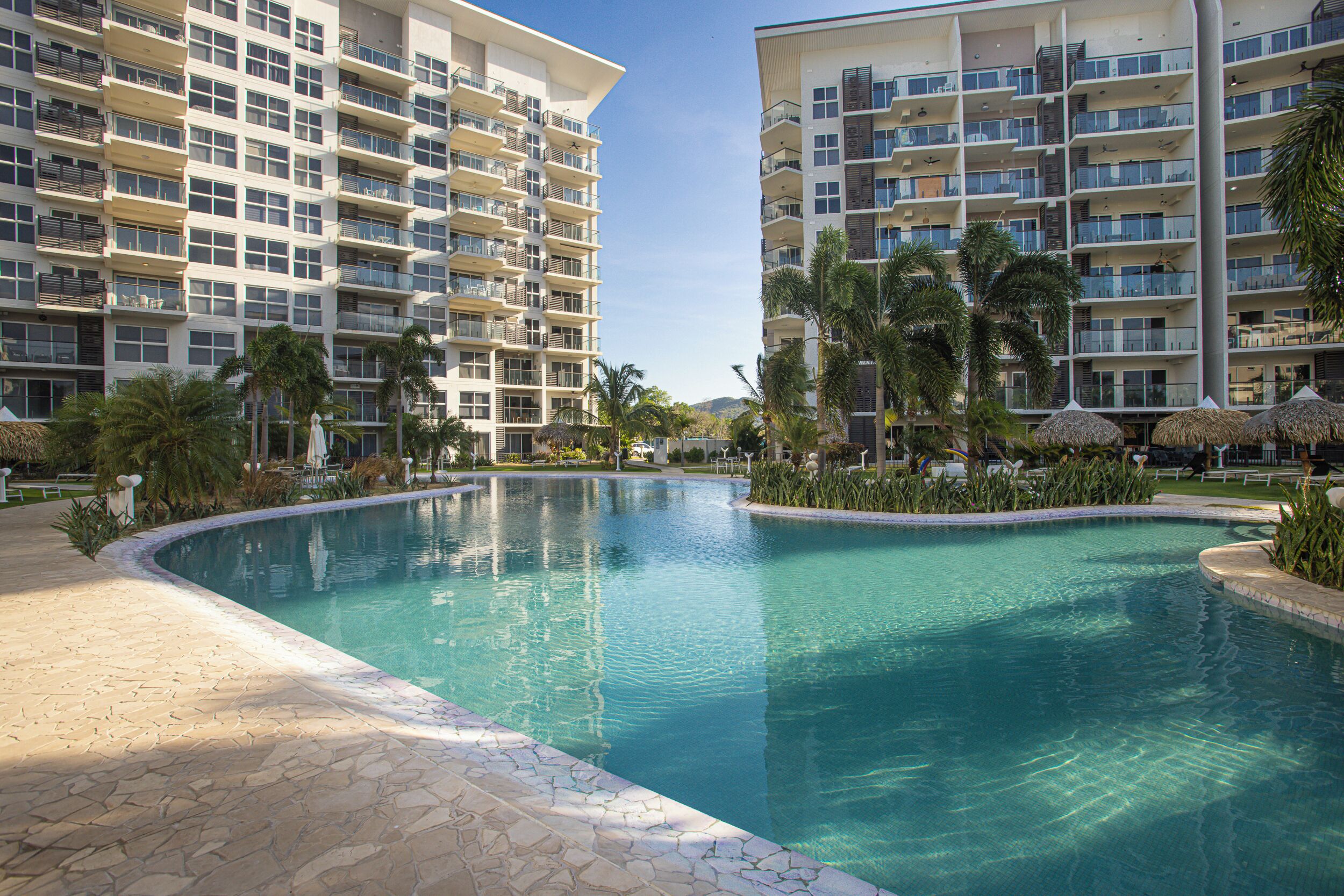 Una piscina rodeada de edificios de varios pisos con balcones, palmeras y un cielo despejado.