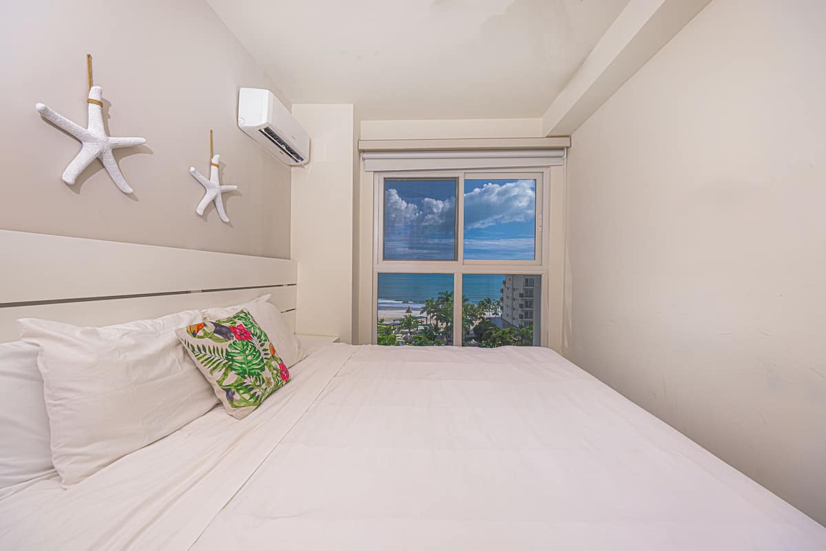 Une chambre à coucher avec un lit, du linge de lit blanc, un coussin coloré, une fenêtre donnant sur la plage et les bâtiments, et deux étoiles de mer décoratives accrochées au mur.