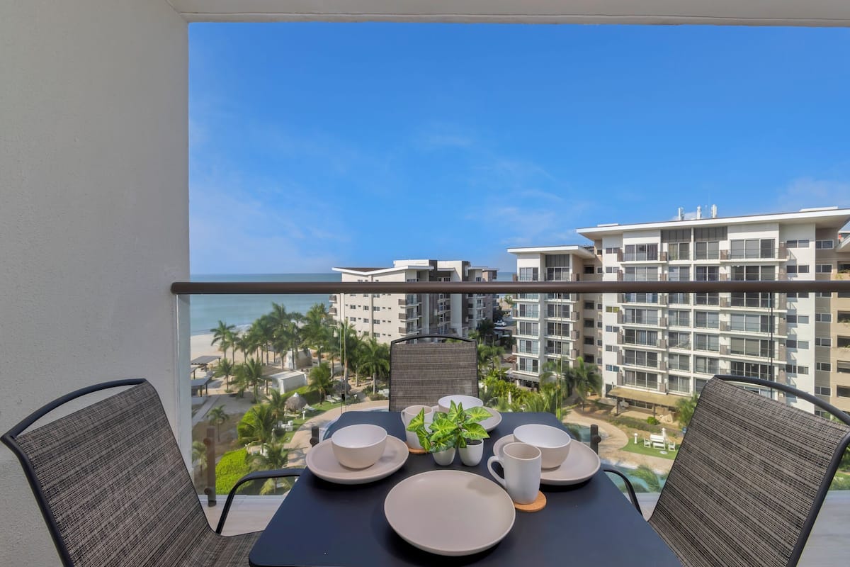 Un balcon avec une table dressée pour deux personnes, donnant sur une zone de villégiature bordée de palmiers et d’une plage.