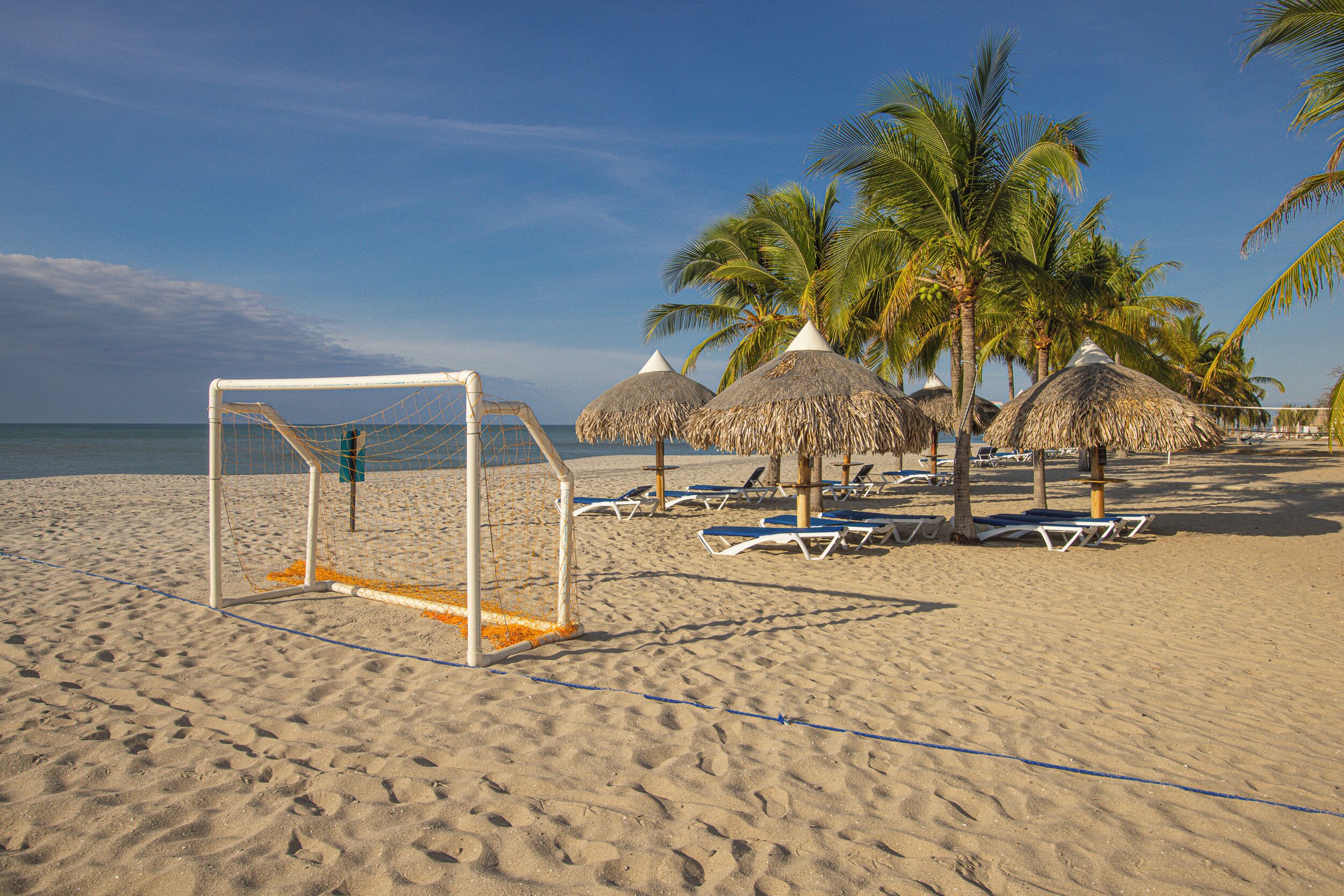 A beach with a soccer goal, palm trees, and sun loungers.