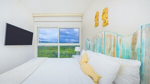 A bedroom with a large bed, a TV mounted on the wall, and a view of greenery through the window.