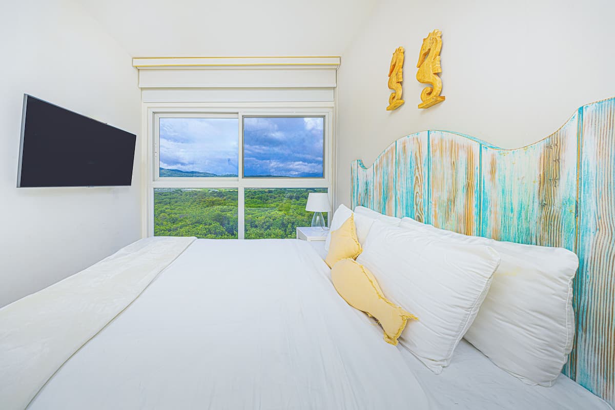 Une chambre avec un grand lit, un téléviseur fixé au mur et une vue sur la verdure par la fenêtre.