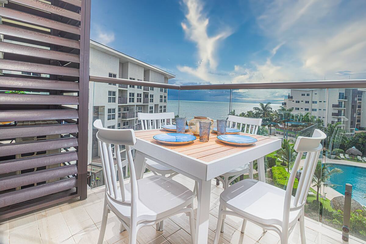 Un balcon avec une table dressée pour deux personnes, donnant sur une piscine et offrant une vue sur l’océan.