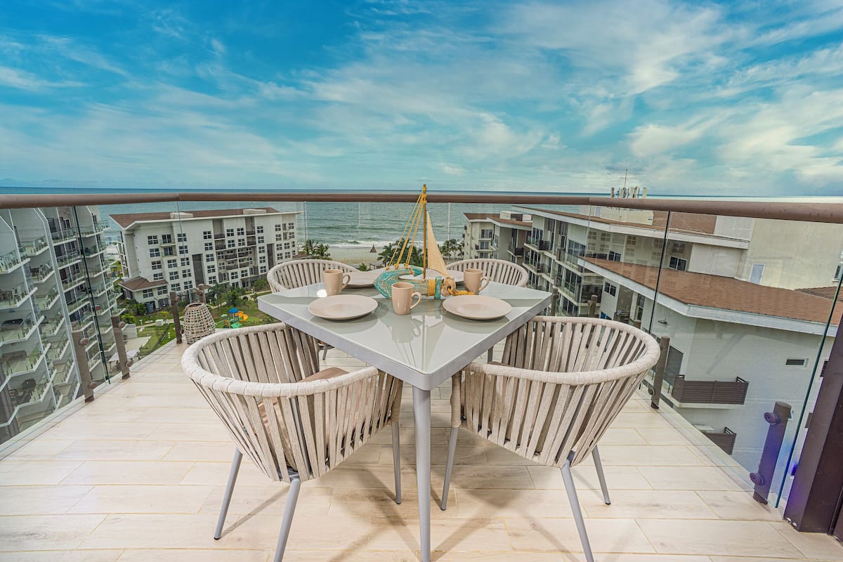 Un balcon avec une table en verre, des chaises en osier et une vue sur l’océan et les bâtiments.