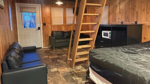 Een houten blokhut met een slaapzolder, een houten ladder, een bank, een magnetron en een deur met ramen.